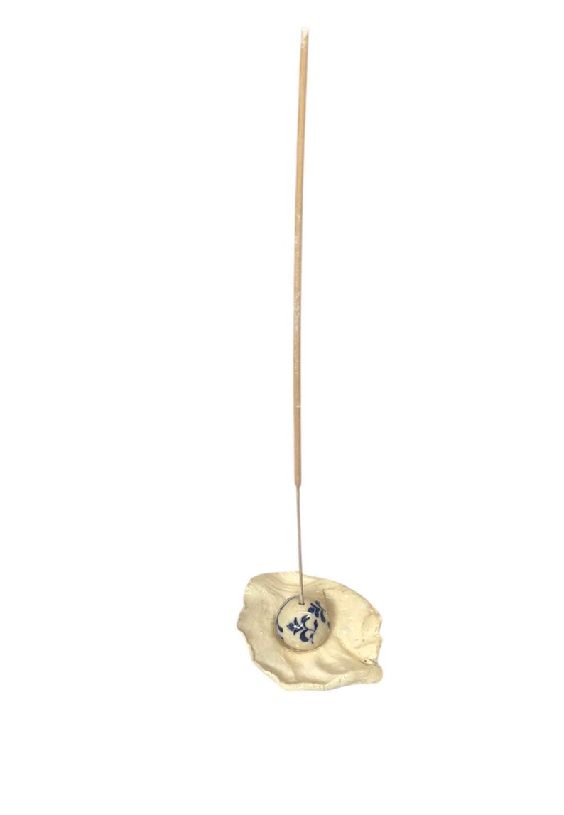 May include: A beige incense holder with a small, round, blue and white floral patterned ceramic ball. A thin, tan incense stick is inserted into the ball, and the holder has a textured, organic shape.