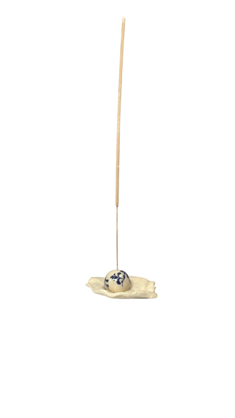 May include: A ceramic incense holder with a cream-colored base and a blue and white patterned sphere. A single incense stick stands upright, ready for use. The holder is designed to catch ash.