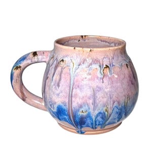 May include: A handmade ceramic mug with a rounded shape and a large handle. The mug features a blend of pink, purple, and blue glaze, with a dripped effect. The mug is suitable for coffee, tea, or other beverages.