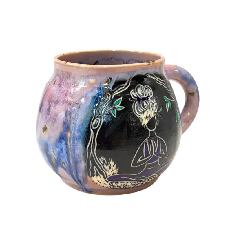 May include: A ceramic mug with a rounded shape and a handle. The mug features a design of a person meditating under a tree, with a black background. The mug has a purple, blue, and pink color scheme.