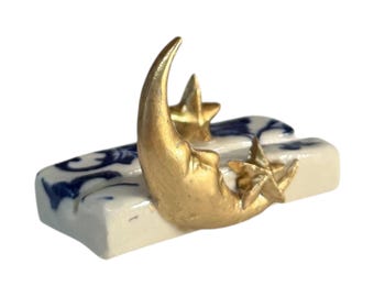 May include: A ceramic rectangular base featuring a white surface with blue floral patterns. Attached to the top is a metallic gold crescent moon figure accompanied by two small gold star shapes, creating a celestial-themed decorative object.