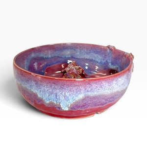 Op de afbeelding: Een ronde keramische kom met een levendig glazuur in tinten magenta, paars en wit. De binnenkant van de kom bevat in het midden een gevormd, bloemachtig keramisch element en een kleine decoratieve uitstulping aan de buitenrand.