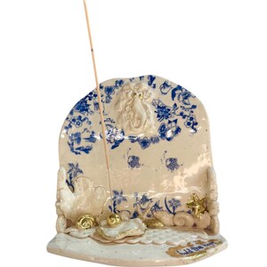 May include: A ceramic incense holder with a cream base and blue floral patterns. The holder features a mermaid figure, gold accents, and the text "hold the hope". An incense stick is inserted.