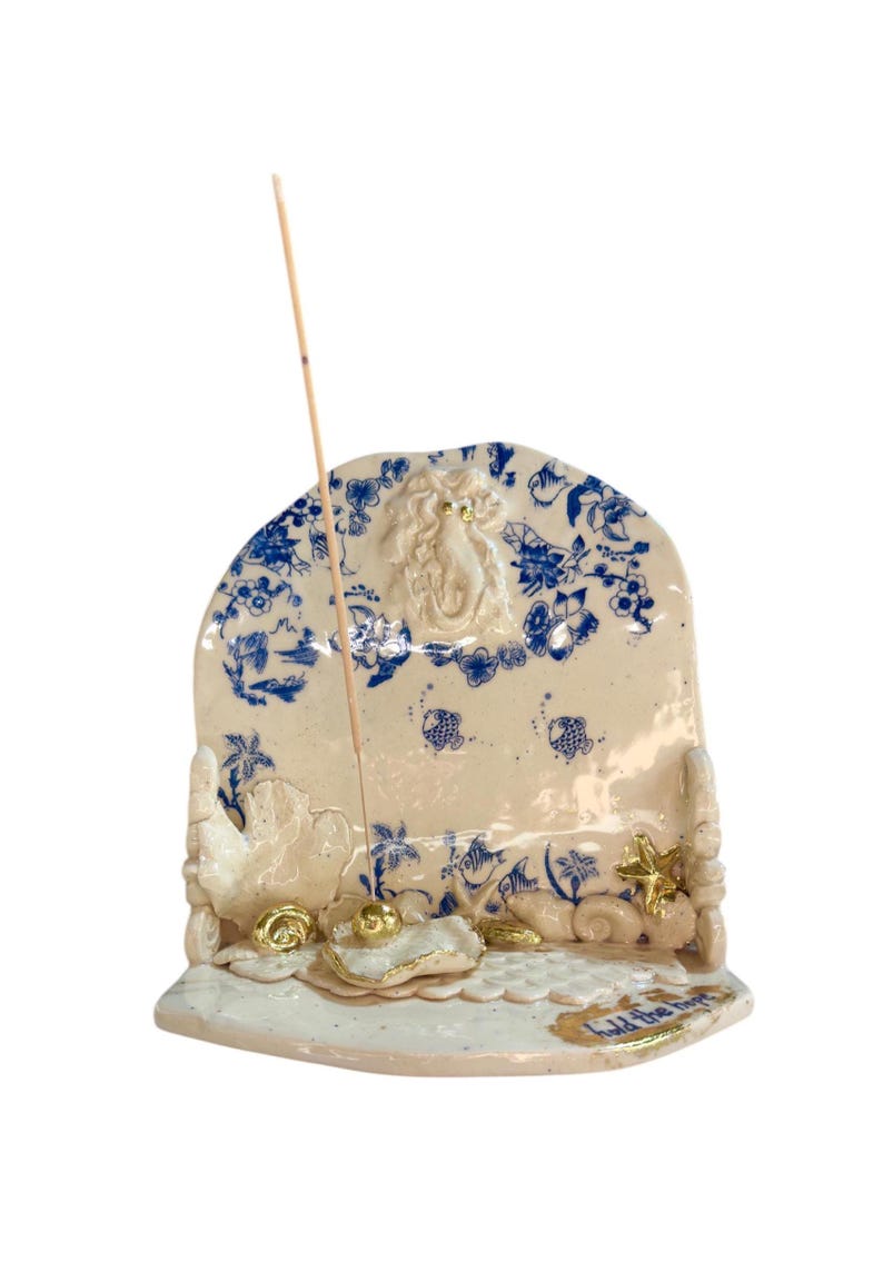 May include: A ceramic incense holder with a cream base and blue floral patterns. The holder features a mermaid figure, gold accents, and the text "hold the hope". An incense stick is inserted.