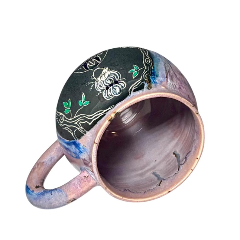 May include: A handmade ceramic mug with a unique floral design. The mug features a black top with white floral and green leaf accents. The body of the mug is a blend of purple, blue, and pink hues. The handle is also a blend of blue and pink.