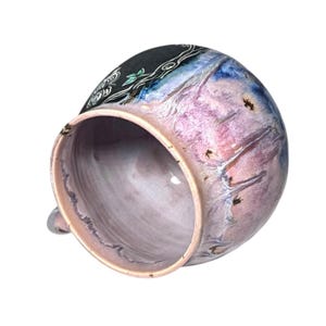 May include: A handmade ceramic mug with a unique design. The mug features a blend of colors, including pink, blue, and black, with a decorative floral pattern. The interior is a soft pink, and the mug has a small handle.