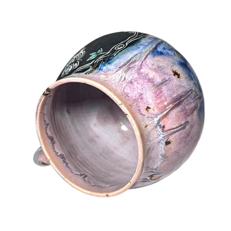 May include: A handmade ceramic mug with a unique design. The mug features a blend of colors, including pink, blue, and black, with a decorative floral pattern. The interior is a soft pink, and the mug has a small handle.