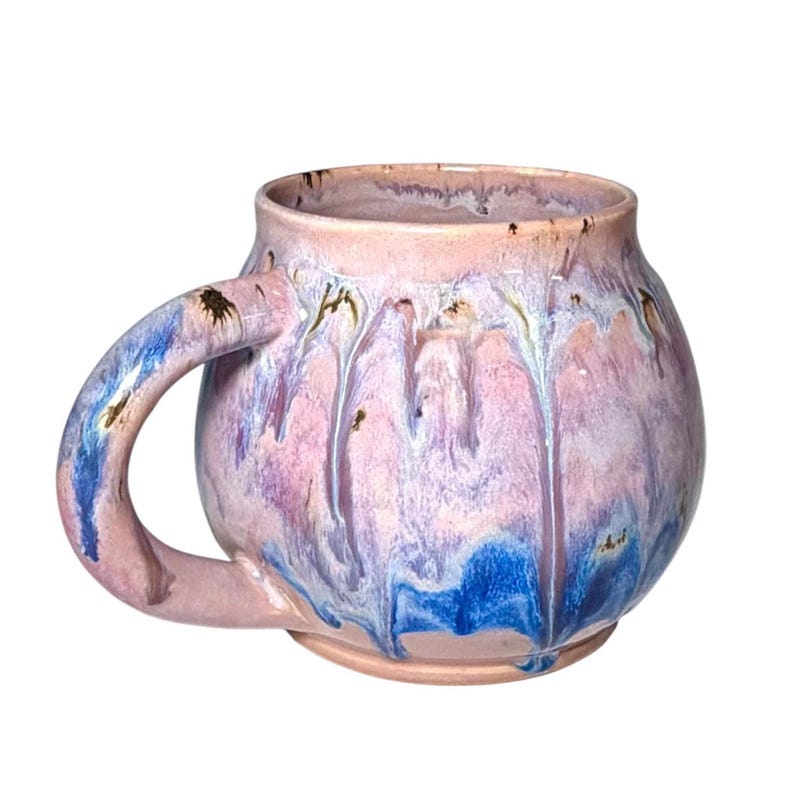 May include: A handmade ceramic mug with a rounded body and a large handle. The mug features a blend of pink, purple, and blue glaze with a dripped effect. The rim and handle have touches of gold.
