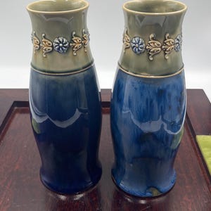 May include: Two ceramic vases with a glossy finish, featuring a deep blue base that transitions into a muted sage green upper section. A decorative band of raised, molded floral and leaf motifs encircles the neck of each vase, adding texture.