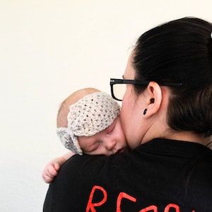 May include: A woman with long black hair wearing glasses and a crocheted hair scrunchie holds a sleeping baby wearing a crocheted headband.