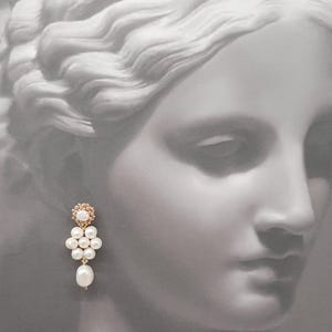May include: A gold-toned earring with a cluster of white pearls and a single dangling pearl. The top of the earring features a small, circular design with a central stone. The background is a grayscale image of a classical sculpture.