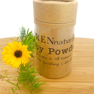 May include: A cylindrical cardboard container of REN natural baby powder with a lid. The container is next to a yellow flower and green foliage. The text on the container reads "Organic & All Natural With Soothing Calendula."
