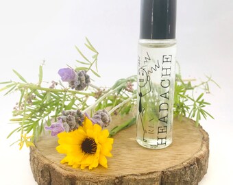 May include: A clear glass roll-on bottle with a black cap, labeled "R.I.A. HEADACHE" and a cartoon figure with a headache. The bottle is on a wooden slice with a yellow flower and sprigs of lavender and greenery.