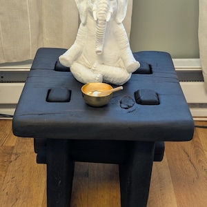 May include: A black wooden stool with a square top and four legs, featuring a white elephant figurine in a seated position and a small gold-colored metal bowl with a wooden striker placed on the surface of the stool.