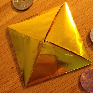 May include: A golden origami creation, folded into a four-pointed star shape. The metallic paper reflects light, creating a shiny effect. The origami is on a wooden surface, with a few coins visible in the background.