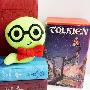 Puede incluir: Un gusano de peluche verde con ojos negros y gafas, que lleva una pajarita roja, está sentado sobre una pila de libros. Los libros se titulan "A Game of Thrones", "A Feast for Crows", "A Dance with Dragons" y "Tolkien".