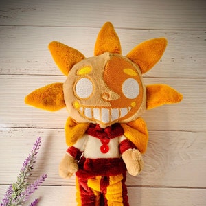 May include: A plush toy shaped like a sun with a smiling face. The sun is wearing a red and yellow jester costume with a white collar and a red button. The toy is standing on a light wood surface.