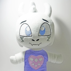 May include: White plush toy with a purple dress and a pink heart with a white symbol on it. The toy has blue eyes and a white horn on its head.