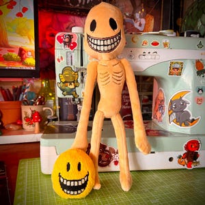 May include: A plush toy with a light orange, elongated humanoid body, long arms, and a round head featuring black oval eyes and a wide, toothy grin. Beside it sits a smaller, round, bright yellow plush with a similar black-eyed, toothy face.