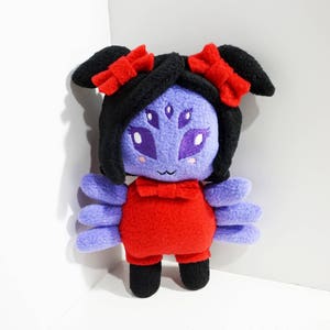 Puede incluir: Un juguete de peluche morado y negro con lazos rojos y un vestido rojo. El juguete tiene tres ojos y una boca sonriente.