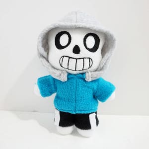 Könnte beinhalten: Ein Plüschtier von Sans aus dem Videospiel Undertale. Das Spielzeug trägt eine blaue Jacke und einen grauen Hoodie. Das Spielzeug hat einen weißen Schädel mit schwarzen Augen und einem schwarzen Lächeln.