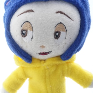 Puede incluir: Mu&ntilde;eco de peluche de un personaje con rostro blanco, ojos bordados y boca roja. El mu&ntilde;eco tiene pelo azul y viste un abrigo amarillo con capucha. Los brazos del mu&ntilde;eco est&aacute;n extendidos, sobre un fondo blanco.