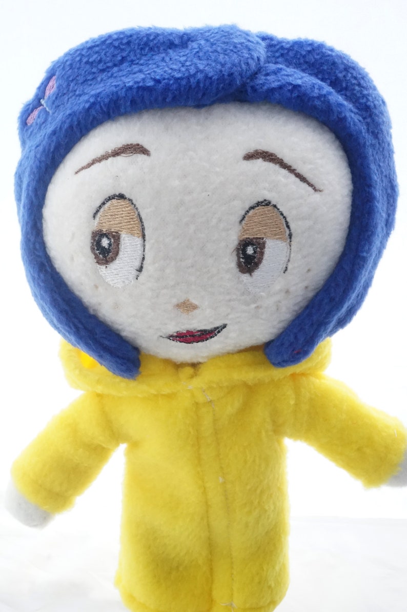 Puede incluir: Mu&ntilde;eco de peluche de un personaje con rostro blanco, ojos bordados y boca roja. El mu&ntilde;eco tiene pelo azul y viste un abrigo amarillo con capucha. Los brazos del mu&ntilde;eco est&aacute;n extendidos, sobre un fondo blanco.