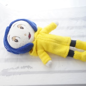 Puede incluir: Mu&ntilde;eca de peluche de un personaje con cabello azul, abrigo amarillo y pantal&oacute;n negro. La mu&ntilde;eca tiene rasgos faciales bordados, incluyendo ojos marrones y una sonrisa roja. La mu&ntilde;eca est&aacute; acostada sobre una superficie blanca.