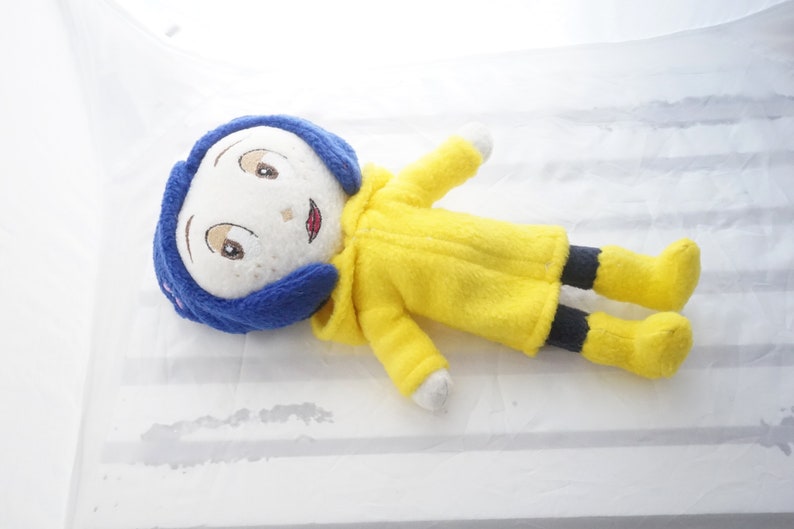 Puede incluir: Mu&ntilde;eca de peluche de un personaje con cabello azul, abrigo amarillo y pantal&oacute;n negro. La mu&ntilde;eca tiene rasgos faciales bordados, incluyendo ojos marrones y una sonrisa roja. La mu&ntilde;eca est&aacute; acostada sobre una superficie blanca.