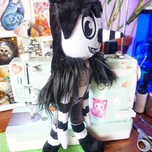 May include: A black and white plush toy with a long black nose and large eyes. The toy is wearing a black hat and a striped shirt. The toy is standing on a green cutting mat.