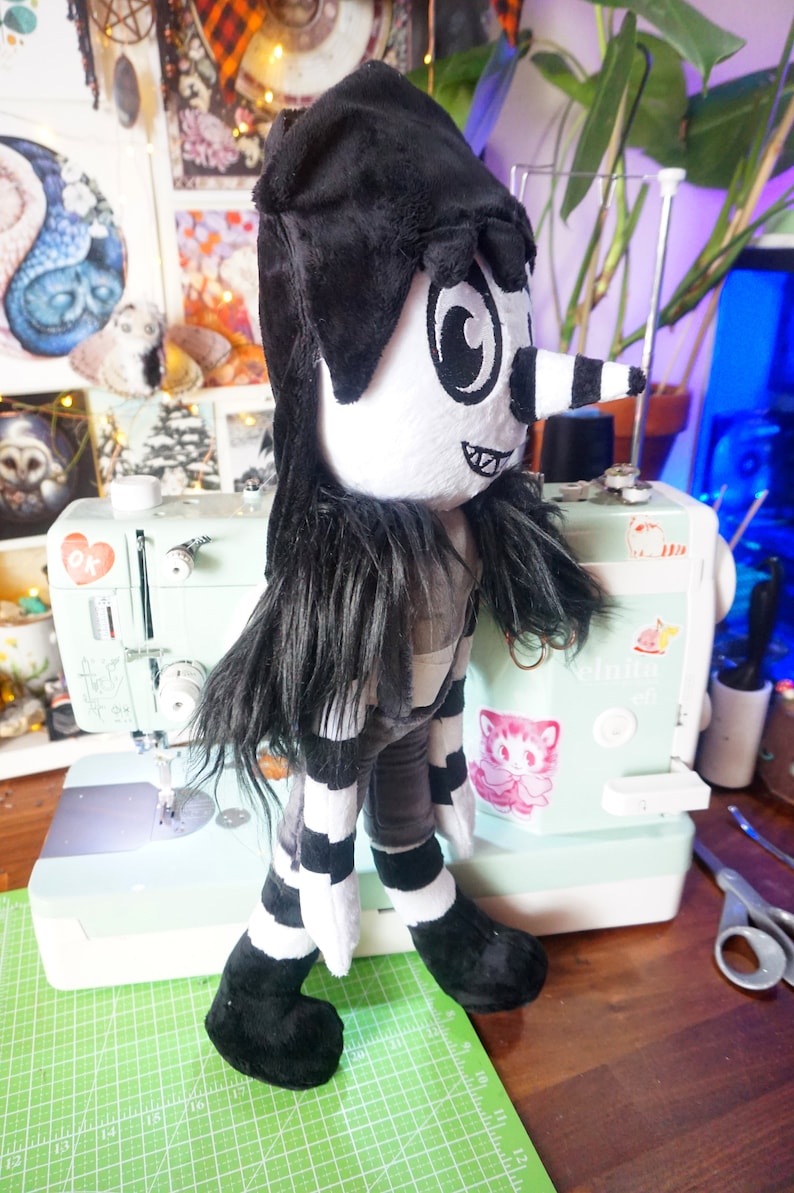 May include: A black and white plush toy with a long black nose and large eyes. The toy is wearing a black hat and a striped shirt. The toy is standing on a green cutting mat.