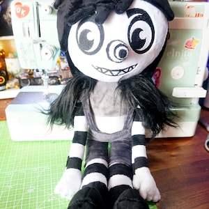 May include: A plush toy of a character with black hair, a white face, and a large, black, one-eyed grin. The toy is wearing a black and white striped outfit and is sitting on a green cutting mat.