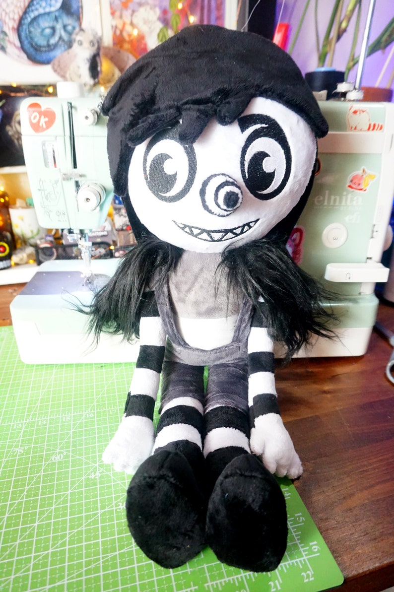 May include: A plush toy of a character with black hair, a white face, and a large, black, one-eyed grin. The toy is wearing a black and white striped outfit and is sitting on a green cutting mat.