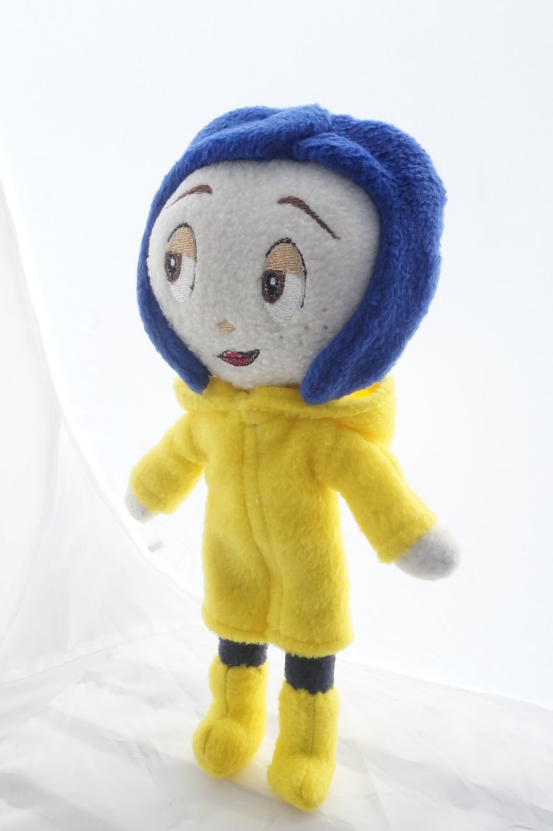 Puede incluir: Mu&ntilde;eca de peluche de un personaje con rostro blanco, ojos bordados y una peque&ntilde;a sonrisa. La mu&ntilde;eca viste una capucha azul y un impermeable amarillo con botas a juego. Los brazos de la mu&ntilde;eca est&aacute;n extendidos, sobre un fondo blanco.