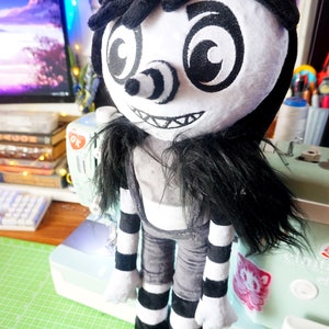 May include: A black and white plush toy with a creepy smile, black hair, and striped trousers. The toy has large, round eyes with black swirls.