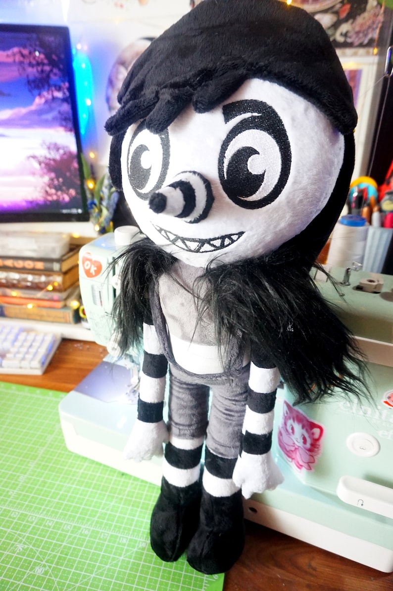 May include: A black and white plush toy with a creepy smile, black hair, and striped trousers. The toy has large, round eyes with black swirls.