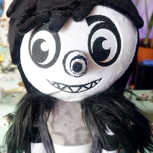 May include: A black and white plush toy with a creepy smile, one eye, and a black hat. The toy has long black hair and is wearing a black and white striped outfit.