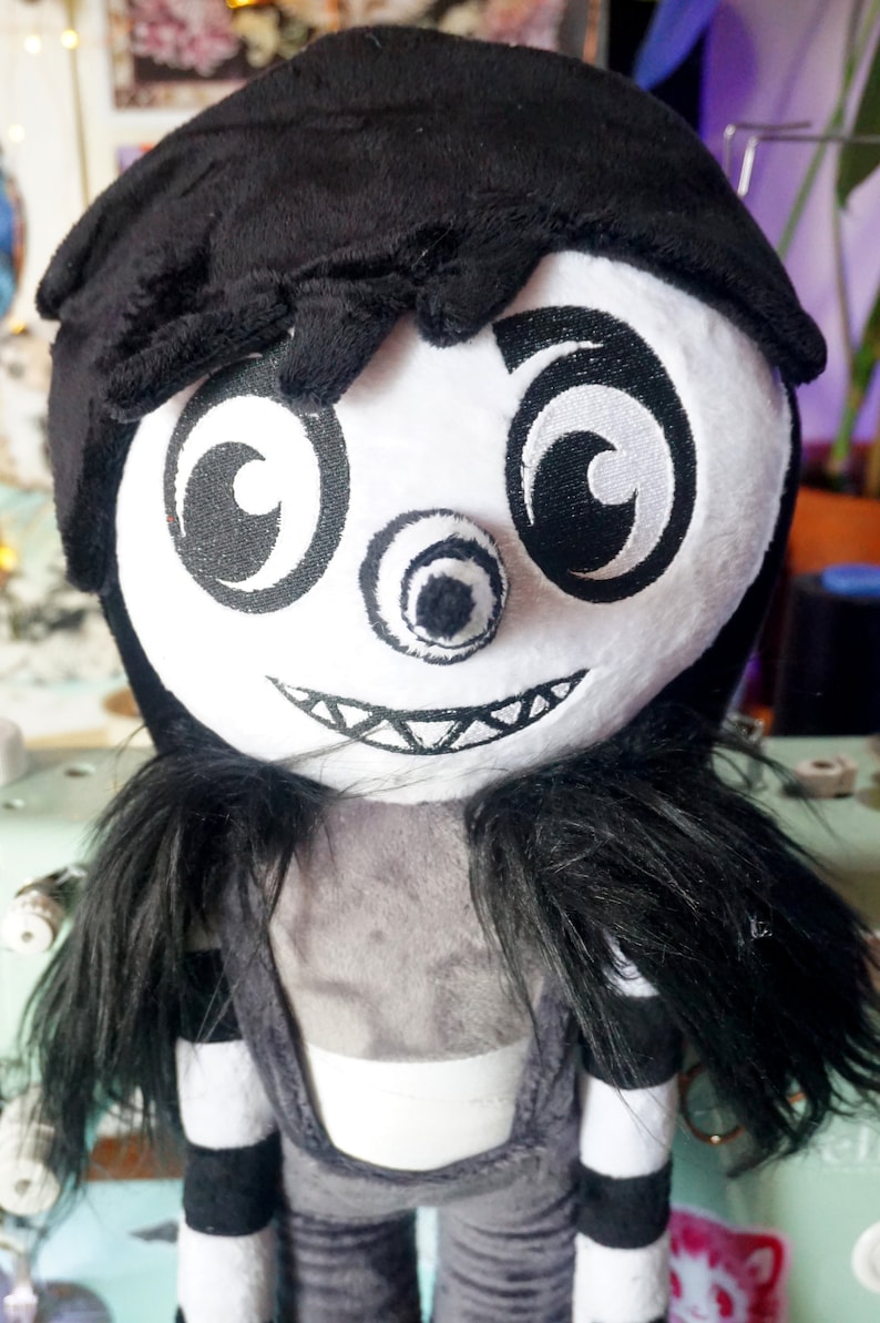 May include: A black and white plush toy with a creepy smile, one eye, and a black hat. The toy has long black hair and is wearing a black and white striped outfit.