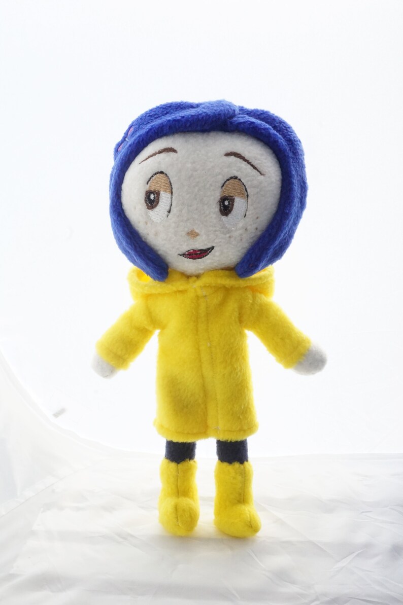 Puede incluir: Mu&ntilde;eca de peluche hecha a mano de un personaje con rostro blanco, ojos bordados y boca roja. La mu&ntilde;eca lleva una peluca azul, un impermeable amarillo con capucha y botas amarillas.