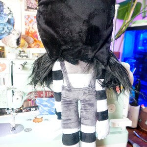 May include: A plush toy with a black and white striped outfit and a black hood covering its head. The toy has long black hair and is standing on a table.