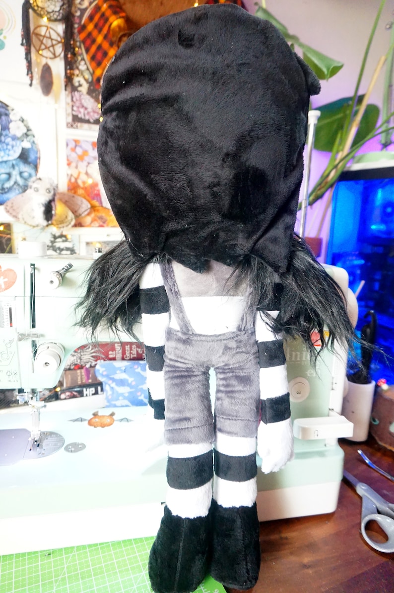 May include: A plush toy with a black and white striped outfit and a black hood covering its head. The toy has long black hair and is standing on a table.
