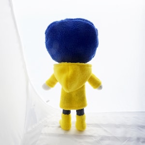 Puede incluir: Mu&ntilde;eco de peluche de un personaje con una gran cabeza azul, que lleva un abrigo con capucha amarillo y botas amarillas. El mu&ntilde;eco se ve desde atr&aacute;s, con las manos blancas y pantalones negros visibles. El fondo es blanco.