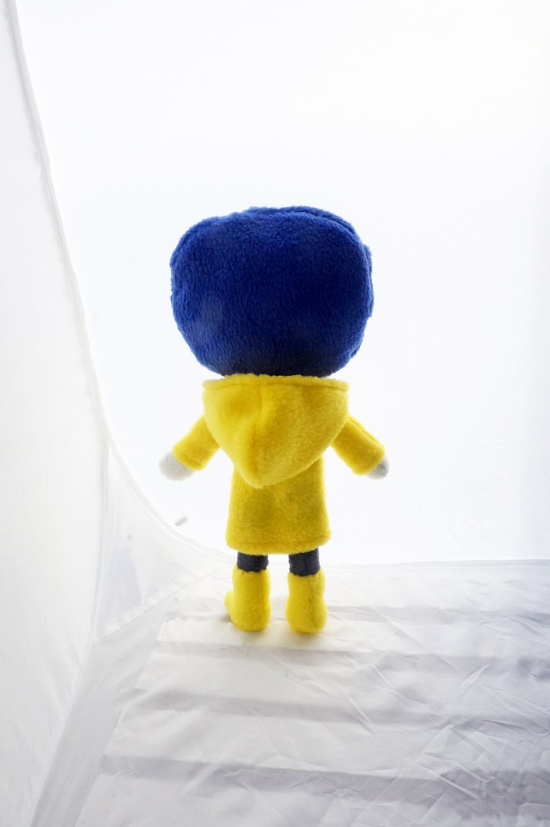 Puede incluir: Mu&ntilde;eco de peluche de un personaje con una gran cabeza azul, que lleva un abrigo con capucha amarillo y botas amarillas. El mu&ntilde;eco se ve desde atr&aacute;s, con las manos blancas y pantalones negros visibles. El fondo es blanco.