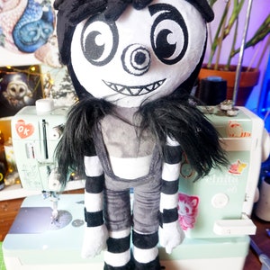 May include: A plush toy of a character with black hair, a white face, and a large, black eye. The toy is wearing a black and white striped outfit and black shoes. The toy is standing on a green cutting mat.
