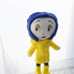 Puede incluir: Mu&ntilde;eca de peluche de un personaje con gorro azul, impermeable amarillo y botas amarillas. La mu&ntilde;eca tiene una cara blanca con rasgos bordados, incluyendo ojos marrones y una boca roja peque&ntilde;a. Fondo blanco.