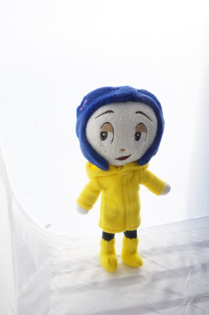 Puede incluir: Mu&ntilde;eca de peluche de un personaje con gorro azul, impermeable amarillo y botas amarillas. La mu&ntilde;eca tiene una cara blanca con rasgos bordados, incluyendo ojos marrones y una boca roja peque&ntilde;a. Fondo blanco.