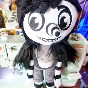 May include: A black and white plush toy with a creepy smile, large eyes, and a spiral nose. The toy is wearing a black and white striped outfit and has long black hair.
