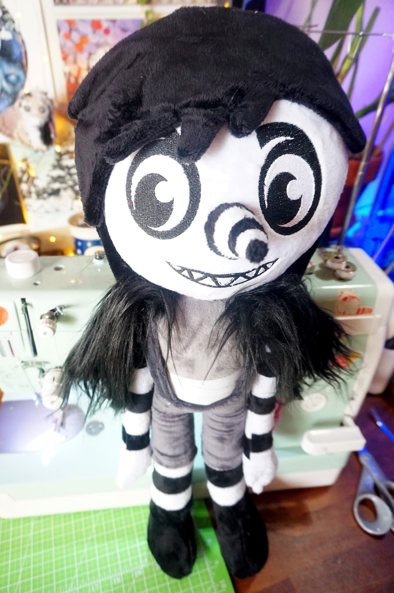 May include: A black and white plush toy with a creepy smile, large eyes, and a spiral nose. The toy is wearing a black and white striped outfit and has long black hair.