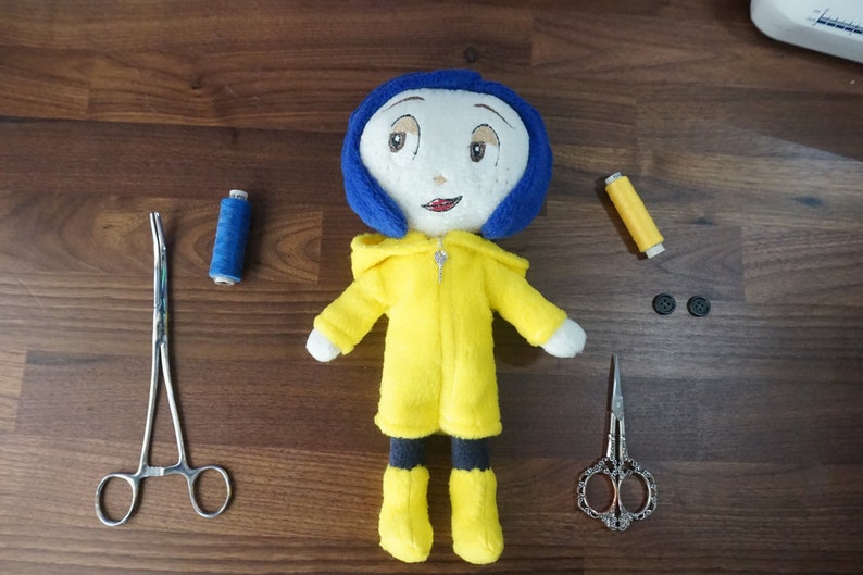 Puede incluir: Una mu&ntilde;eca de peluche hecha a mano con cabello azul y un abrigo amarillo, que se asemeja a un personaje. Suministros de costura, incluyendo tijeras, hilo y botones, est&aacute;n dispuestos alrededor de la mu&ntilde;eca sobre una superficie de madera. La mu&ntilde;eca mide aproximadamente 20 cm de altura.