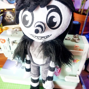 May include: A black and white plush toy with a creepy smile, large eyes, and a long black wig. The toy is wearing a black and white striped outfit and black shoes.